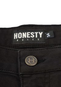 Schwarze Hosen aus strukturiertem Stoff. Merkmale sind ein Etikett mit der Aufschrift "EHRLICHKEIT REGIERT" sowie ein Metallknopf in Diamantform mit dem "R"-Logo.