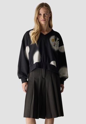 Woman wearing a black oversized sweatshirt with white abstract spots and a small cartoon bird, paired with black pleated pinstripe culottes.