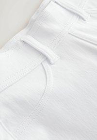 Gros plan sur un tissu en denim blanc montrant une poche, une passants de ceinture et des coutures détaillées sur un jean blanc.