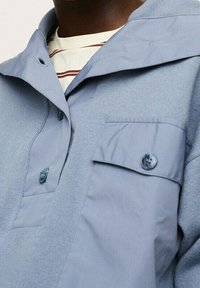 Ingrandimento di una persona che indossa una camicia azzurro chiaro con bottoni, dotata di una tasca frontale e colletto, sopra una camicia bianca con strisce rosse.