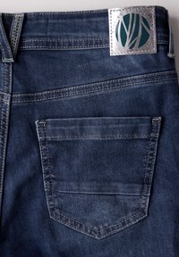 Los vaqueros de mezclilla presentan un lavado oscuro con costuras en contraste, un bolsillo trasero y una etiqueta plateada con un logo verde en la cinturilla.