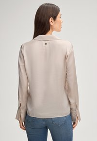 Zijde beige blouse met een kraag en lange mouwen, met een knoopaccent op de rug en gespleten manchetten voor extra detail.