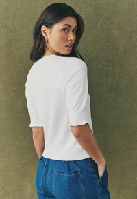 Pull blanc à manches courtes et froncées ; ourlet côtelé et texture douce. Porté avec un pantalon en denim bleu, mettant en valeur un look décontracté.