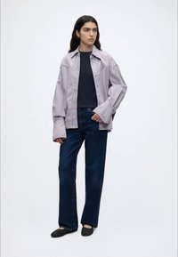 Γυναίκα όρθια που φορά ανοιχτό μωβ υπερμεγέθη πουκάμισο με κουμπιά, μαύρο μπλουζάκι, σκούρα μπλε τζιν και μαύρα υφασμένα slip-on παπούτσια.