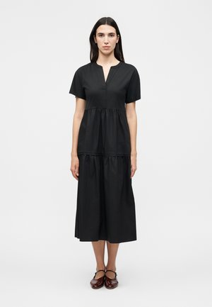 Žena s rovnými tmavými vlasmi, oblečená v čiernych midi šatách s krátkym rukávom a stupňovitou sukňou, s hnedými topánkami Mary Jane, stojaca na bielom pozadí.