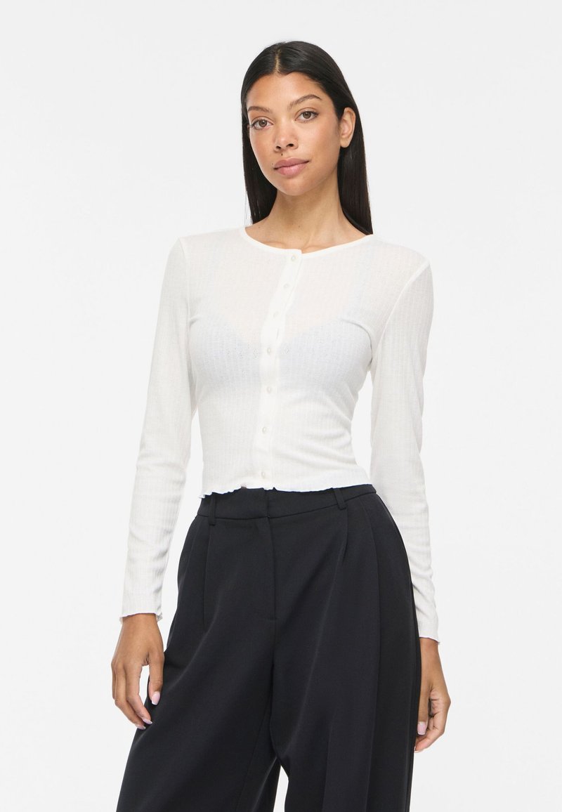 Femme aux cheveux raides foncés portant un haut blanc à manches longues boutonné et un pantalon noir taille haute à jambes larges sur fond blanc.