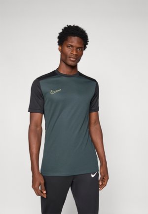 Bărbat îmbrăcat cu un tricou athletic Nike de culoare verde închis și negru și pantaloni Nike negri, stând în fața unui fundal simplu de gri deschis.