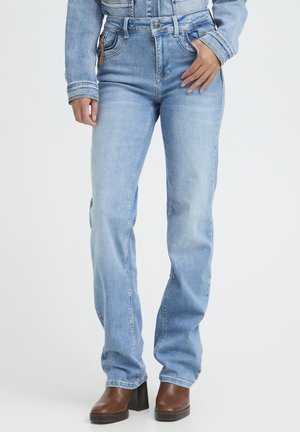 Person iført lyseblå jeans med høj talje og brune blokhælede støvler, med den ene hånd hvilende på hoften.