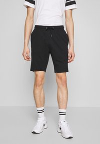 Shorts de sport noirs avec une taille élastique et un cordon de serrage, accompagnés d'une chemise blanche avec des rayures noires et des baskets blanches.