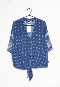 Blouse bleue à manches courtes avec des motifs floraux blancs, décolletée en V, avec un nœud à l'avant, suspendue à un cintre noir sur fond blanc.