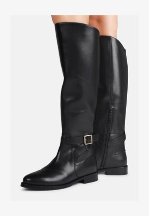 Une paire de bottes en cuir noir, montantes jusqu'au genou, avec fermeture éclair sur le côté et boucle décorative, portées sur des jambes nues devant un fond uni.