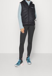 Schwarze gesteppte Weste mit Kapuze, hellblaues Langarmshirt, schwarze Leggings und farbenfrohe Sportschuhe mit blauen Schnürsenkeln.