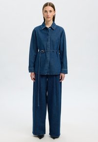 Giacca in denim e pantaloni a gamba larga di colore blu scuro. La giacca ha un colletto, vita con cintura e polsini. Texture liscia con dettagli di cucitura a contrasto.