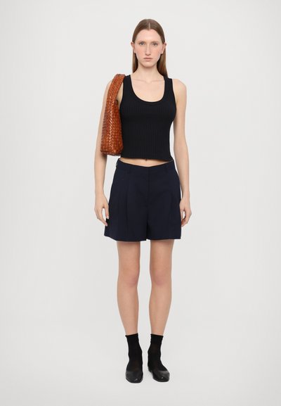 Schwarzes geripptes Cropped Tanktop, marineblaue taillierte Shorts und eine braune geflochtene Handtasche. Model trägt schwarze Socken und Schuhe mit Blockabsatz.