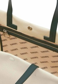 Sac fourre-tout en toile avec des poignées en simili cuir vert foncé. L'intérieur comprend une poche zippée et un logo de la marque répété sur une doublure beige clair.