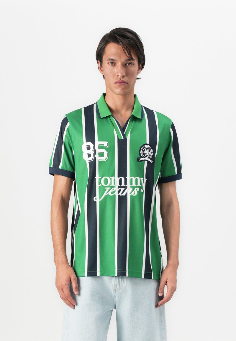 Hombre vistiendo una camiseta polo de rayas verticales en verde, azul marino y blanco con los logos "85" y "tommy jeans", combinada con jeans de color azul claro, de pie contra un fondo blanco.