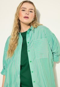 Camisa a rayas verde y blanca con un bolsillo, con un diseño de botones y mangas cortas, superpuesta sobre una camiseta verde sólida.