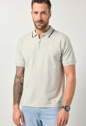 Uomo con capelli castani corti e barba che indossa una polo a maniche corte grigio chiaro con cerniera, jeans blu chiaro, orologio marrone e un tatuaggio dettagliato sull'avambraccio sinistro.