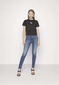 Zwarte katoenen t-shirt met een wit logo, gecombineerd met donkerblauwe skinny jeans en zwarte strap sandaaltjes. Eenvoudig, nauwsluitend ontwerp.