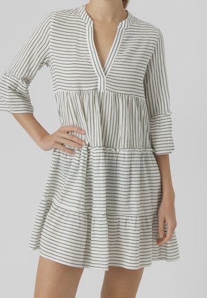 Woman wearing white knee-length dress with thin black horizontal stripes and 3/4 sleeves, posing with one hand on hip.