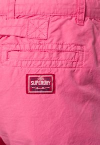 Tissu rose avec rabat de poche arrière et patch rectangulaire rouge et blanc de la marque Superdry cousu sous la poche.