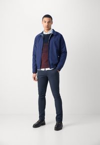 Chaqueta ligera azul marino con cuello alzado, superpuesta a un suéter texturizado burdeos, combinada con pantalones oscuros y mocasines negros.