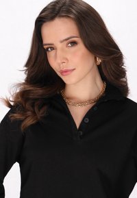 Tricou polo negru cu guler și trei butoane, având o textură fină. Colier din lanț auriu și cercei cercuri aurii ca accesorii notabile.