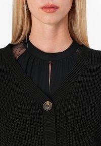 Mujer rubia vistiendo un cárdigan negro texturizado con un botón de concha de tortuga sobre una blusa negra con paneles de encaje en los hombros.