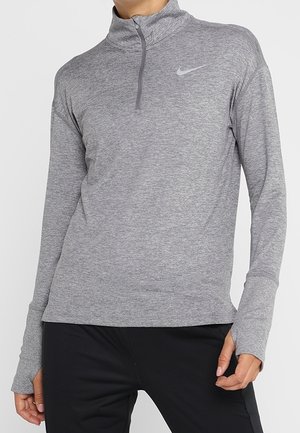 Frau trägt ein hellgraues Nike-Oberteil mit langem Arm und Viertel-Reißverschluss mit Daumenlöchern, kombiniert mit schwarzen Hosen.