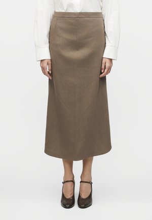 Donna che indossa una gonna midi marrone a trapezio, una camicetta bianca a maniche lunghe e scarpe Mary Jane nere con tacco, in piedi davanti a uno sfondo semplice.