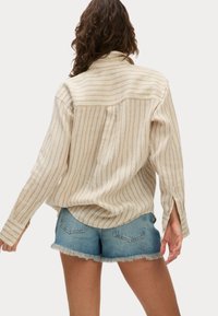 Camicia oversize a righe beige chiaro con maniche lunghe, abbinata a pantaloni shorts in denim blu sfilacciati. La camicia presenta una spalla posteriore e polsini con bottone.