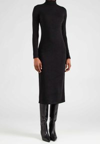 Frau in einem langärmligen schwarzen Rollkragen-Midi-Kleid, kombiniert mit schwarzen knielangen Lederstiefeln, steht vor einem schlichten Hintergrund.