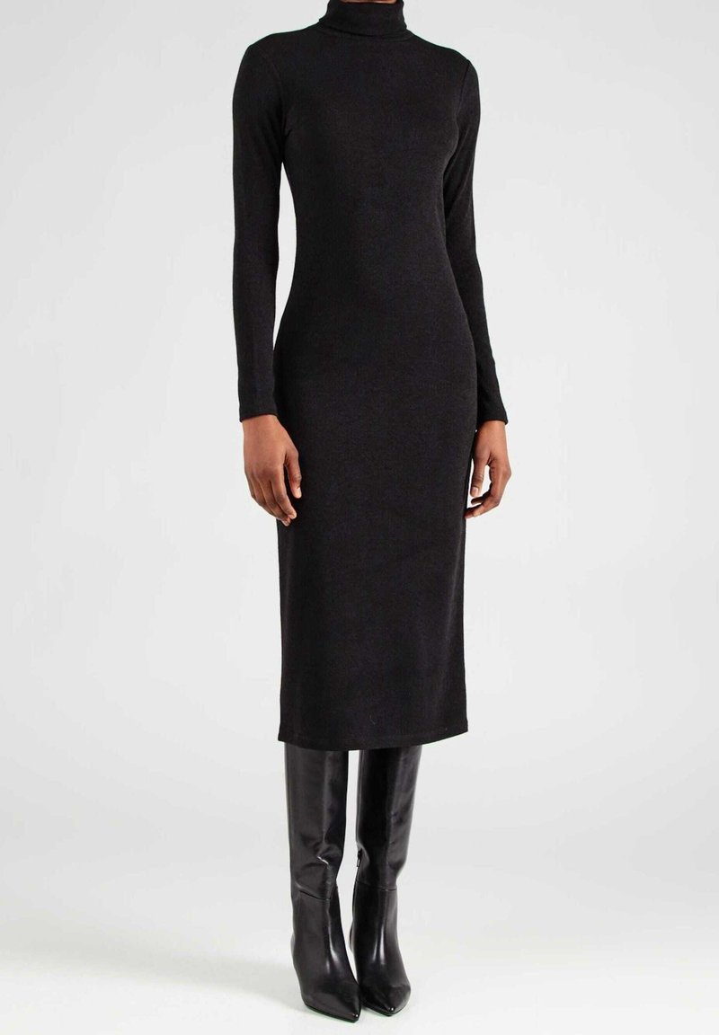 Frau in einem langärmligen schwarzen Rollkragen-Midi-Kleid, kombiniert mit schwarzen knielangen Lederstiefeln, steht vor einem schlichten Hintergrund.