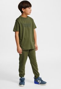 Zielona bawełniana koszulka z teksturowanym logo, w połączeniu z zielonymi spodniami dresowymi. Dziecko ma na sobie niebieskie buty sportowe z białymi akcentami.