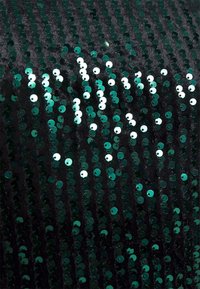 Černá látka pokrytá uspořádáním lesklých zelených a stříbrných flitrů, vytvářející texturovaný vzor s mírným leskem.