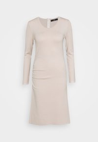 Dlouhý béžový šaty z jemné látky, mají kulatý výstřih a decentní boční řasení, končí těsně nad kolenem s rovným střihem.