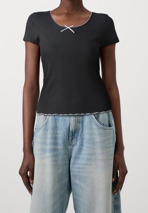 Czarna bluzka na krótki rękaw o prążkowanej fakturze, okrągłym dekolcie, białym wykończeniu i dekoracyjnym krzyżowym detalem na środku. Połączona z jasnoniebieskimi dżinsami.