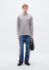 Γκρι μακρυμάνικο πουκάμισο με μικρό λογότυπο, συνδυασμένο με μπλε τζιν τζιν. Αξεσουάρ περιλαμβάνουν μαύρη δερμάτινη τσάντα και μαύρα παπούτσια.