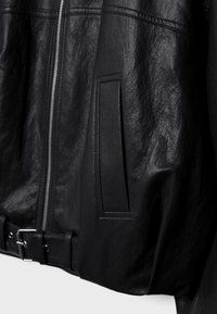 Musta nahkatakki, jossa pystysuuntainen vetoketju, sivutasku ja säädettävä vyö vyötäröllä, joka korostaa tekstuurillista materiaalia ja ompelutaitoja.