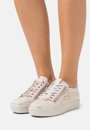 Füße, die hellbeige Plateausneaker mit glänzenden Satin-Schnürsenkeln und silbernen Seit-Reißverschlüssen auf weißem Hintergrund tragen.