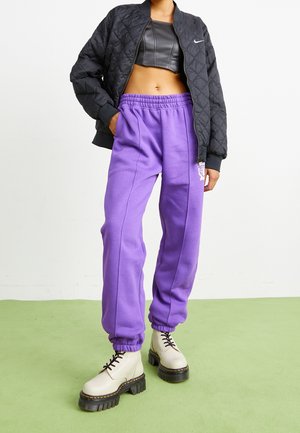 Personne portant un pantalon de survêtement violet, des bottes blanches épaisses à lacets, une veste matelassée noire et un crop top noir, debout sur un sol vert contre un mur blanc.