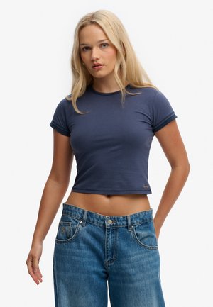 Mujer con cabello largo y rubio, vistiendo una camiseta corta ajustada azul marino y jeans azules sueltos contra un fondo liso.