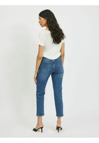 Jeans de denim azul com cintura ajustada e corte reto, acompanhados por uma t-shirt branca e sapatos de salto pretos, vistos de trás.