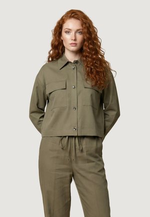 Joven mujer con pelo rojo rizado y largo, vistiendo una chaqueta verde oliva abotonada y pantalones a juego con cordón, de pie con las manos detrás de la espalda.