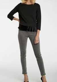 Svart tröja med 3/4-ärmar och veckad nederkant, kombinerad med grå slim-fit byxor. Tröjan har diskreta axelknappar och en mjuk textur.