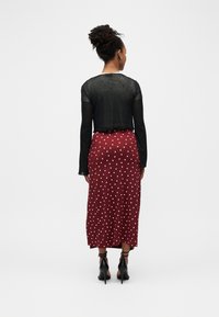 Femme aux cheveux bouclés attachés en chignon, portant un haut noir transparent à manches longues, une jupe midi rouge avec des motifs de cœurs blancs, et des talons hauts noirs, debout.