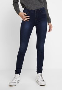 Smalbyxor i mörkblå denim med hög midja, fem fickor och subtila sömnadsdetaljer, i kombination med vita högklackade sneakers.