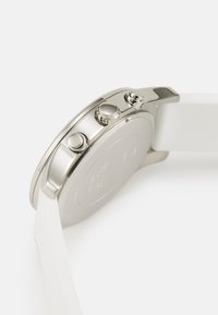 Silverfärgad klocka med rund klocka, krona och tryckknappar. Har ett vitt gummiband och en slät, polerad metallfinish. Baksidan är blank.