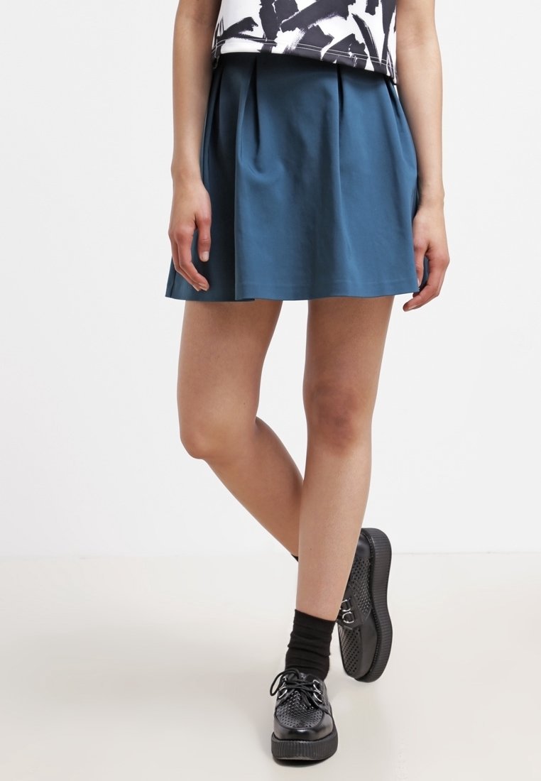 Una gonna plissettata color turchese realizzata in tessuto liscio, caratterizzata da un design svasato. Abbinata a sneakers nere con texture e calze scure.