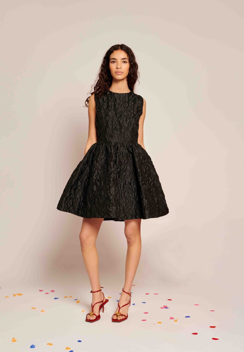 Mulher com cabelo longo e encaracolado, vestindo um vestido preto sem mangas e texturizado e sandálias de salto alto vermelhas, de pé num chão coberto de confetes.